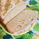 An iced loaf cake decorated with diagonal sprinklings of ground pistachios and ground almonds sits on a narrow yellow platter. The end slice is cut to show the interior texture of the cake, which is light-colored and lightly flaked with ground pistachios, ground almonds, and cardamom. The narrow yellow platter rests on a vintage napkin with a rough watercolor-like print of green and yellow strawberries, arranged on a green tablecloth.