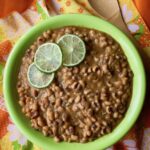 A green bowl of black-eyed peas in a ruddy orange and coconut sauce flavored with ginger and five-spice powder. The peas are garnished with thin slices of lime. The bowl rests on a orange, yellow, and green napkin printed with daisies and an orange tablecloth. A wooden serving spoon is to the right of the bowl.
