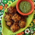 Spherical vegetable fritters made with radishes, carrots, and leeks in an oval-shaped lemongrass-green dish are photographed from above. In the dish with the fritters is a small orange bowl containing a bright green herb & yogurt dipping sauce. The dish of fritters and sauce sits on a vintage napkin printed with a design of white, green, and orange daisies on a brown background.