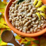 A large orange bowl is photographed containing Great Northern beans cooked with onion, garlic, bay, and lemon. Wedges of lemon and a bay leaf are tucked into the beans as garnishes. The bowl rests on vintage napkins printed with large yellow daisies with brown centers. A wooden serving spoon with a round bowl lies below the bowl of beans.