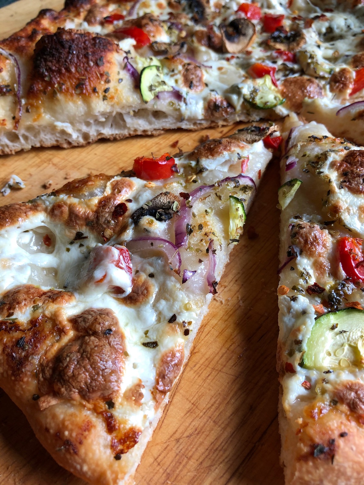A pizza topped with roasted garlic white pizza sauce, fresh mozzarella, red peppers, mushrooms, zucchini, and red onions is photographed from a low angle lying on a light-colored wood cutting board. A slice has been moved back from the pie so that the development of small bubbles in the layer of crust under the toppings can be seen.