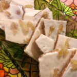 A long green glass dish sits on a psychedelic vintage napkin with a floral pattern in shades of green, pink, orange, and yellow. The dish holds shingled pieces of candy brittle topped with white chocolate and crystallized ginger.